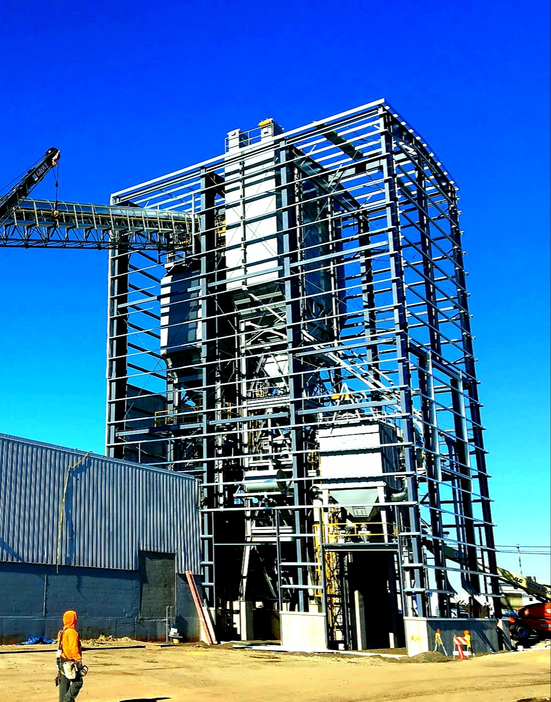 Tall industrial steel lattice structure under construction with internal tanks, a worker in an orange jacket giving scale, and a Grove crane on site