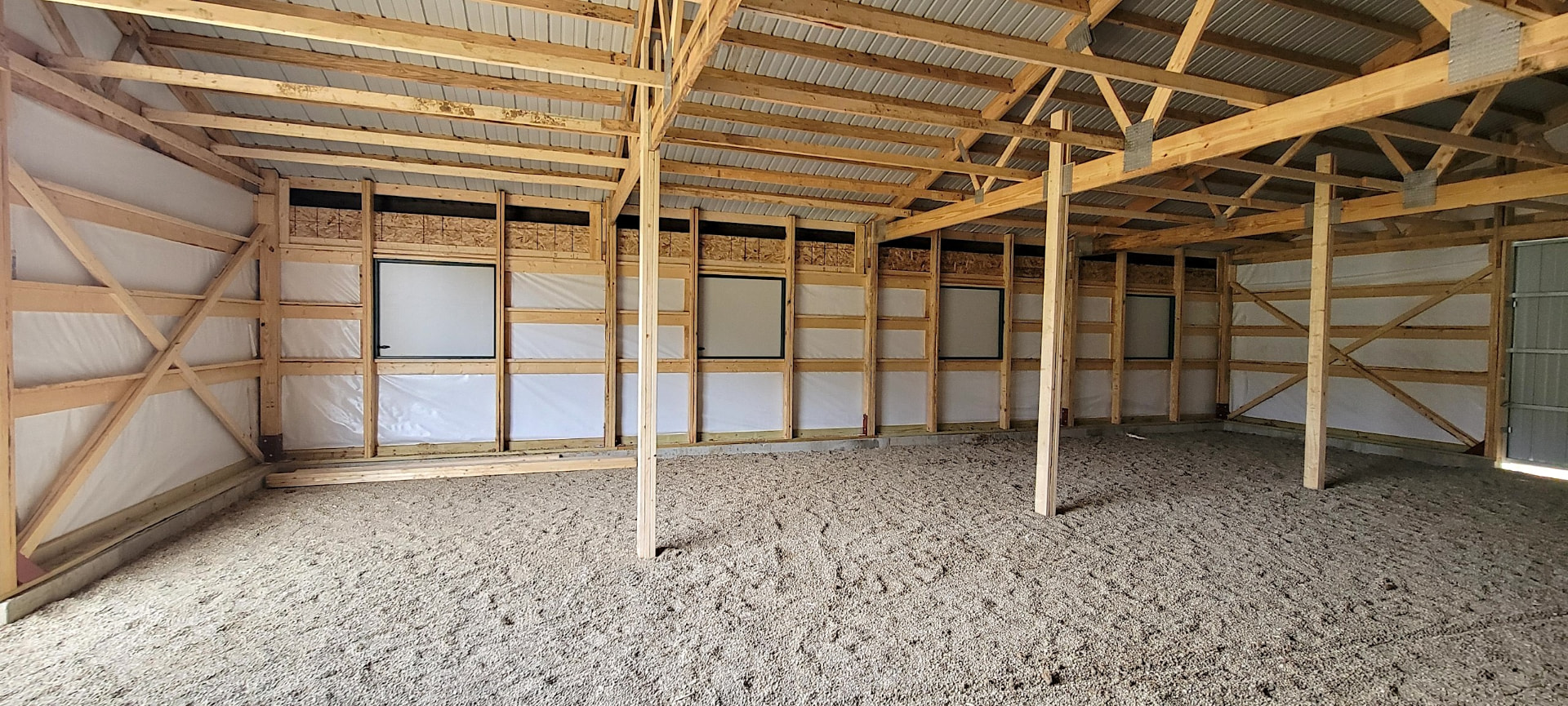 Interior of a large post-frame agriculture building with timber framing, galvanized truss gusset plates, white vapor barrier walls, and a gravel floor